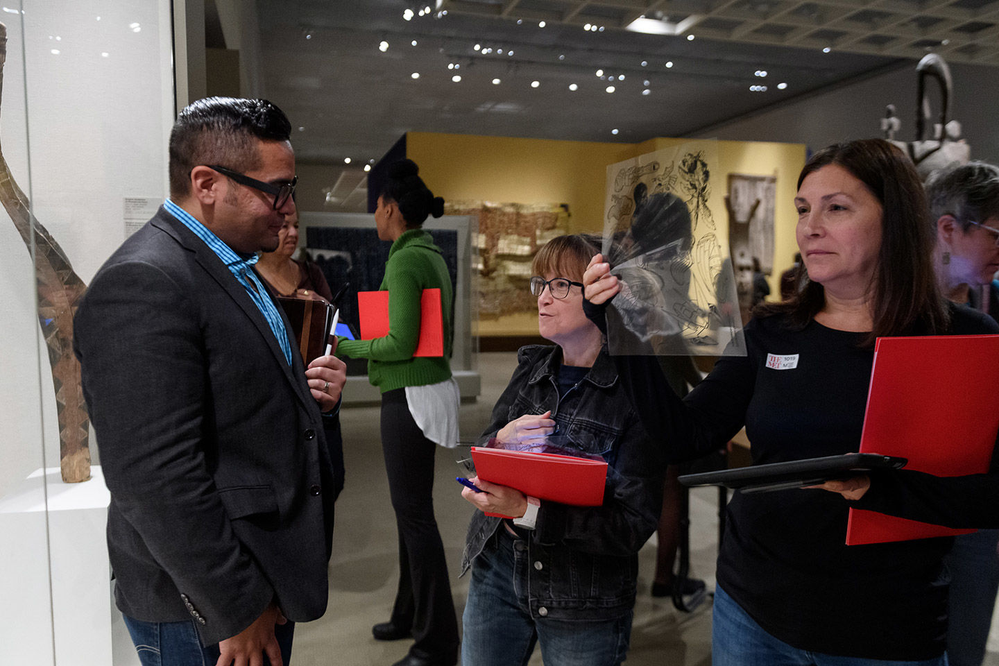 Two women, holding bright red folders, stand before a gallery guide within a gallery space; one converses with the guide while the other holds up a transparency with outlined illustrations; other adults converse in the background